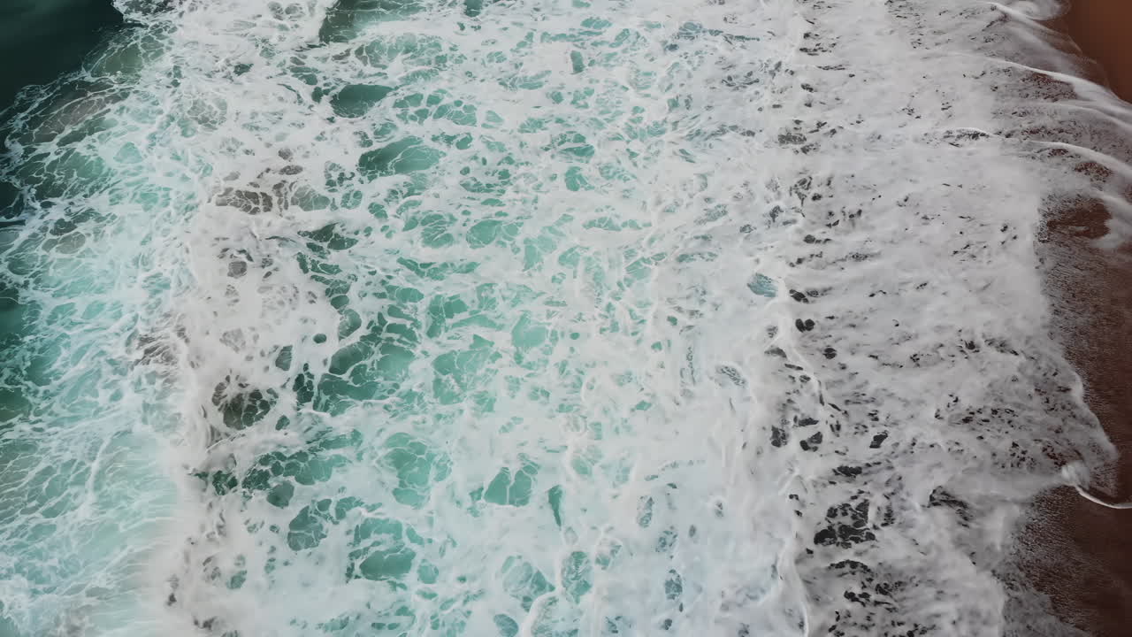 Gentle waves crash onto a sandy beach, creating foamy patterns as the tide rolls in. The sun shines brightly, reflecting on the water's surface, enhancing the tranquil atmosphere