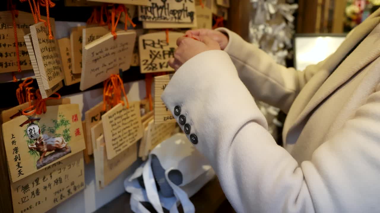 白衣を着た日本人女性が、神社で木製の絵馬を掛ける。