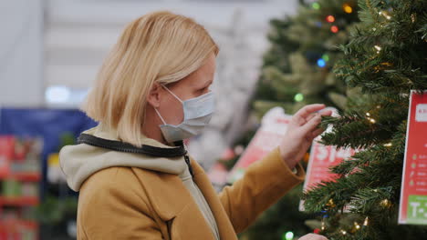 una mujer con una máscara protectora elige un árbol de navidad en la tienda. compras seguras para las vacaciones de invierno