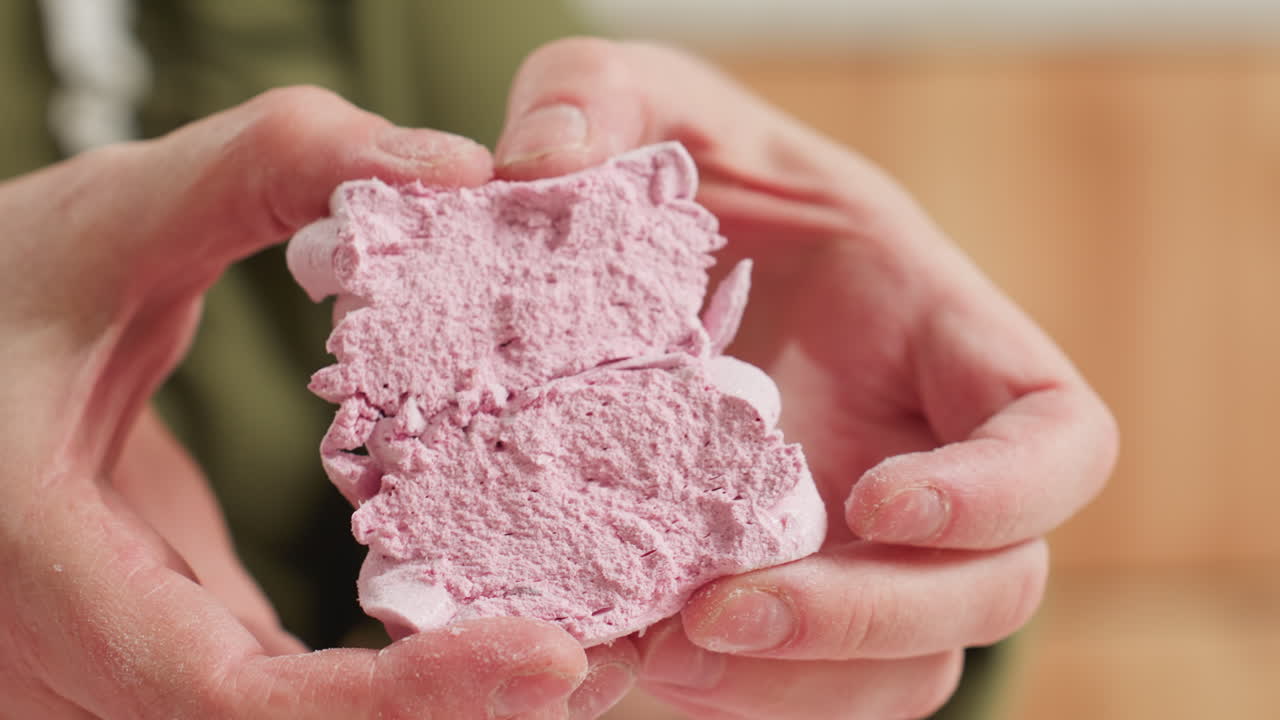 Close up of person gently pressing open pink cupcake showing soft airy texture with playful motion in warm kitchen setting with blurred background