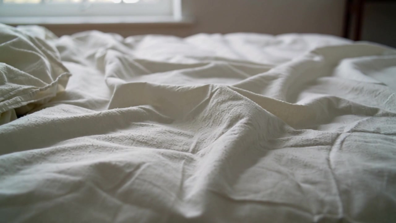 Memindahkan seprai krem yang kusut dari kasur yang sedang dirapikan di dekat jendela kamar tidur, memperlihatkan bantal dan bangku.