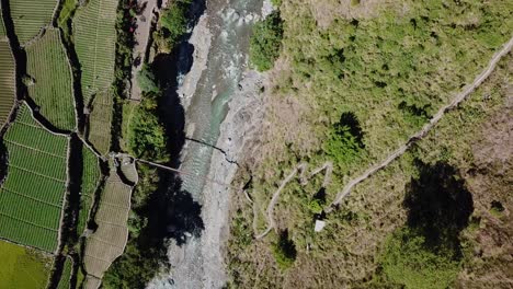 metal-suspension-bridge-spanning-over-river-leading-to-trails-farms-farmers-rice-patties-gardens-trails-on-sunny-day-Top-down-bird's-eye-view