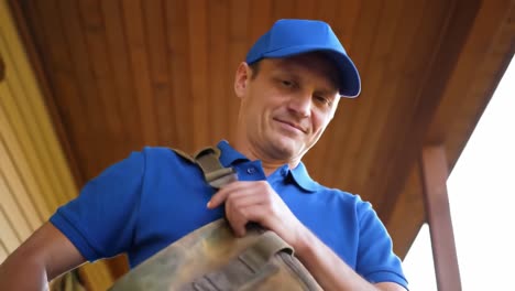 a man in a blue polo shirt and cap smiles while holding a bag at a wooden entrance. sunlight streams in, creating a warm atmosphere as he prepares to head out.