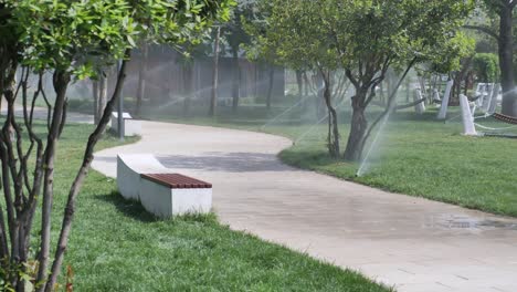 a stone pathway in a park with green grass and trees on either side. there is a bench and a sprinkler system in the grass.