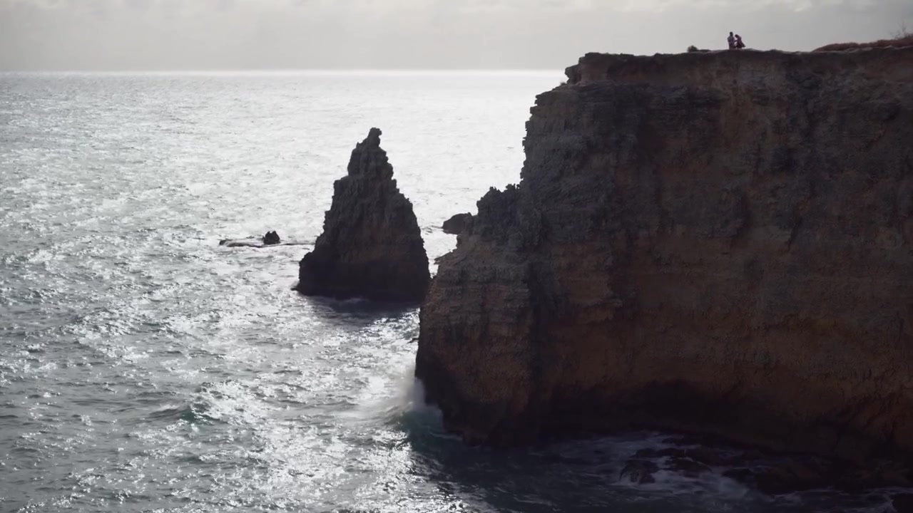 Drone shot of sea waves splashing against rocky cliffs and natural rock arches along the coast on a sunny day.