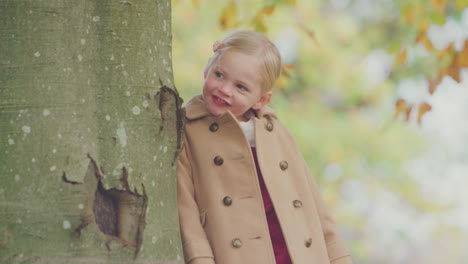 young girl looking around trunk of autumn tree playing hide and seek in garden