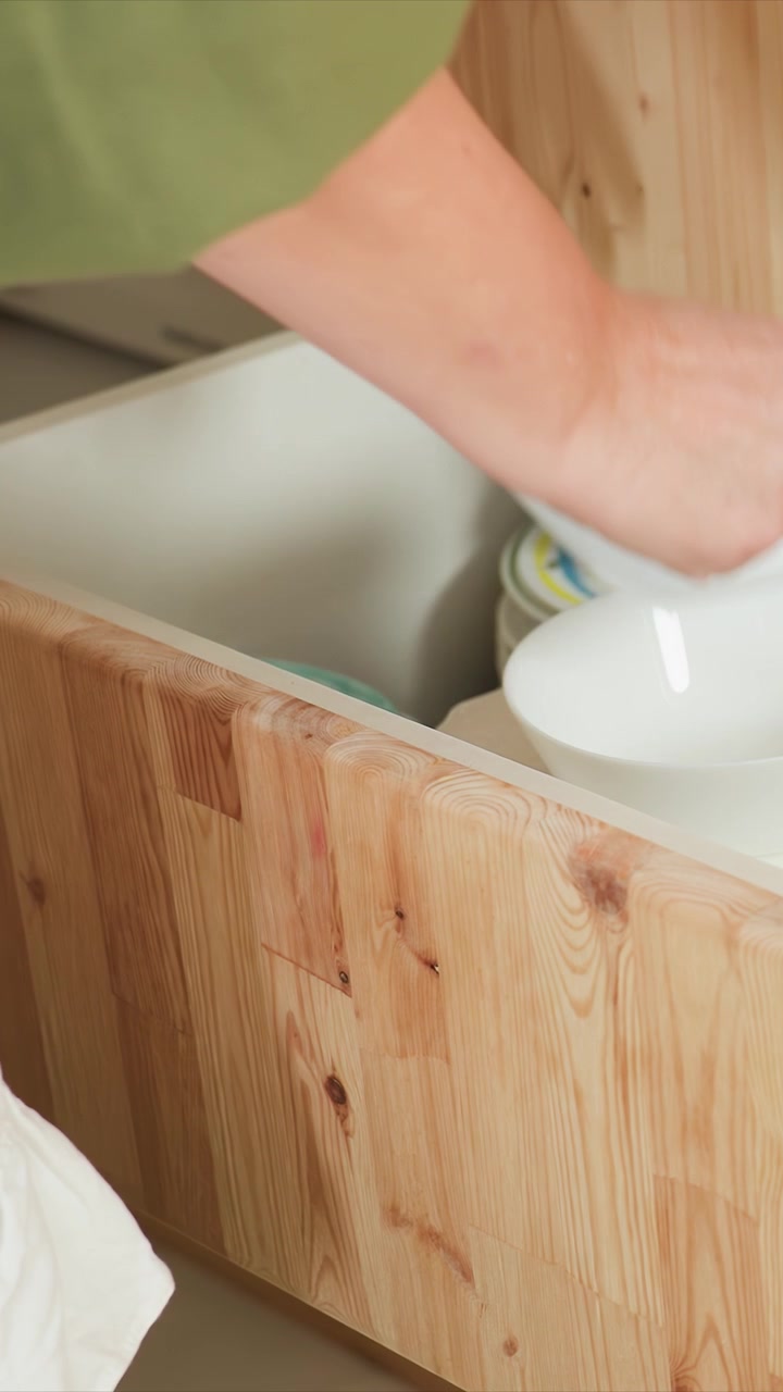 Woman in green top interacts with wooden kitchen cabinets. Scene features close up views of hands handling dishes. Swaying piece of fabric adds subtle movement to domestic setting
