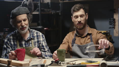 portrait shot of two cheerful father and son carpenters talking and drinking coffee in workshop