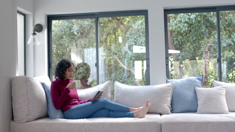 Biracial-woman-sitting-on-sofa-using-tablet-and-drinking-coffee-at-home,-slow-motion,-copy-space