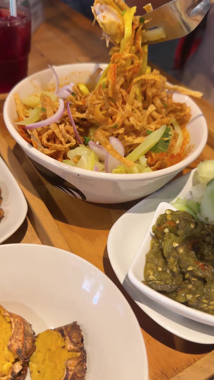 Seseorang mengaduk mi kari Khao Soi dengan sendok di meja kayu yang dilengkapi hidangan pendamping.