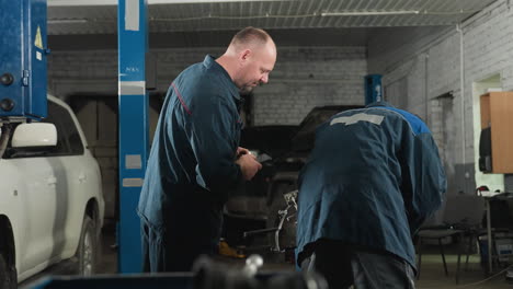 mechanical engineers in a workshop, one intently working on an engine with a wrench, back to the camera, while his colleague uses his phone, surrounded by mechanical equipment