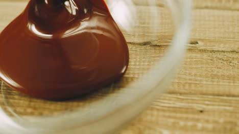 Macro-of-melted-chocolate-pouring-into-transparent-bowl-on-wooden-table.