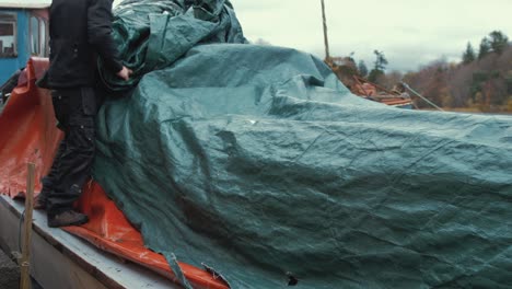 young man unties waterproof cover of wooden boat before starting restoration maintenance