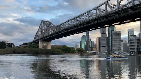 static shot riverside view capturing a citycats passenger ferry boat on the river, cruising across and under the famous heritage story bridge in brisbane city, queensland, australia