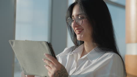 beautiful smiling woman with glasses gently pushes hair back while holding tablet, standing by window in sunlit modern building with urban view and soft natural light enhancing her features