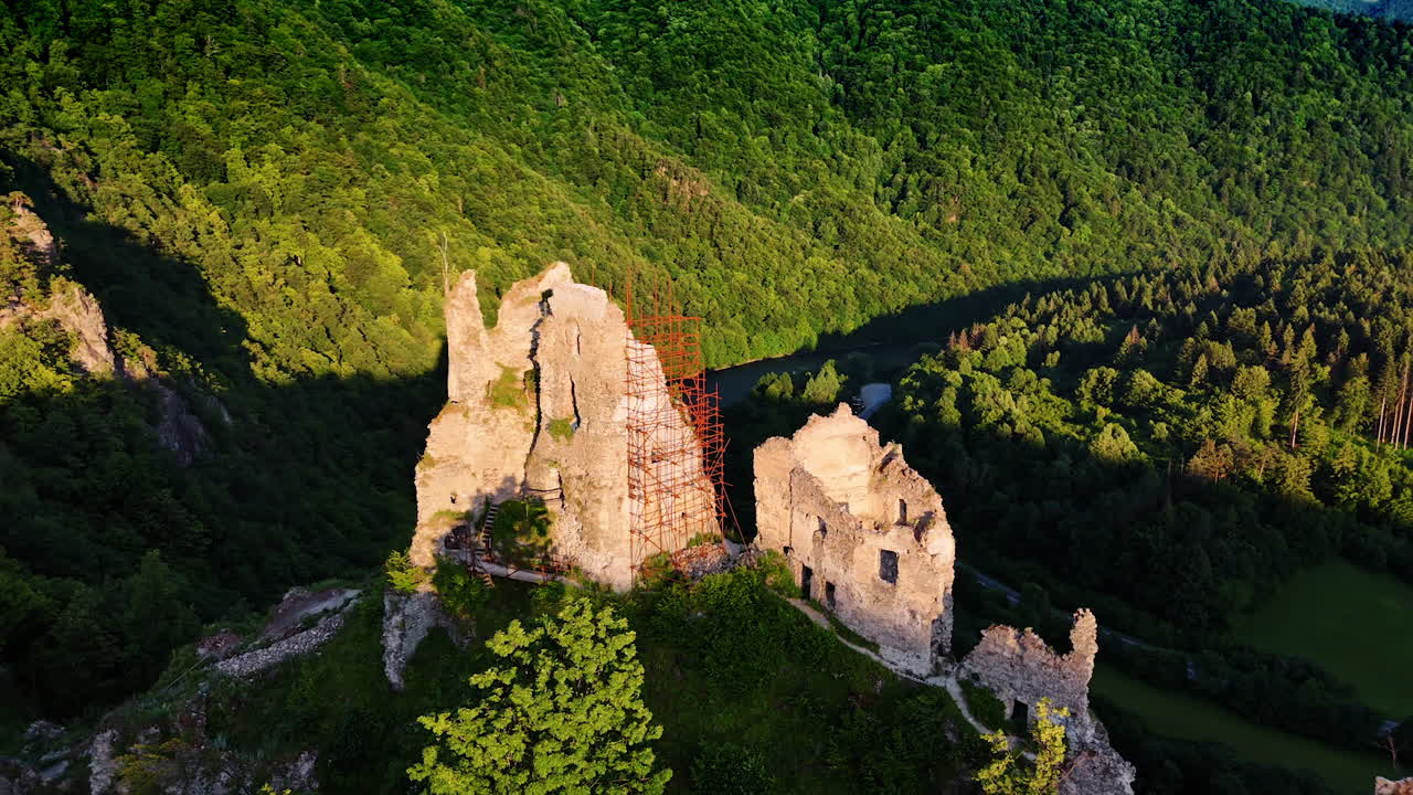 Ancient castle ruins with the scaffolding at one wall. Wooded slopes of the mountains and river at the foot at backdrop. Aerial view.