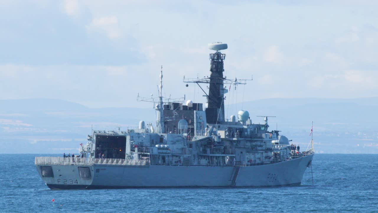 A military vessel steadily sails across calm blue waters near Elie, Fife, Scotland, under daylight with a stable, wide shot and clear sky