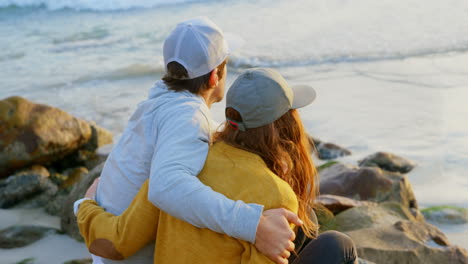 Rückansicht-Eines-Jungen-Kaukasischen-Paares,-Das-Auf-Einem-Felsen-Sitzt-Und-Am-Strand-Auf-Das-Meer-Blickt,-4k