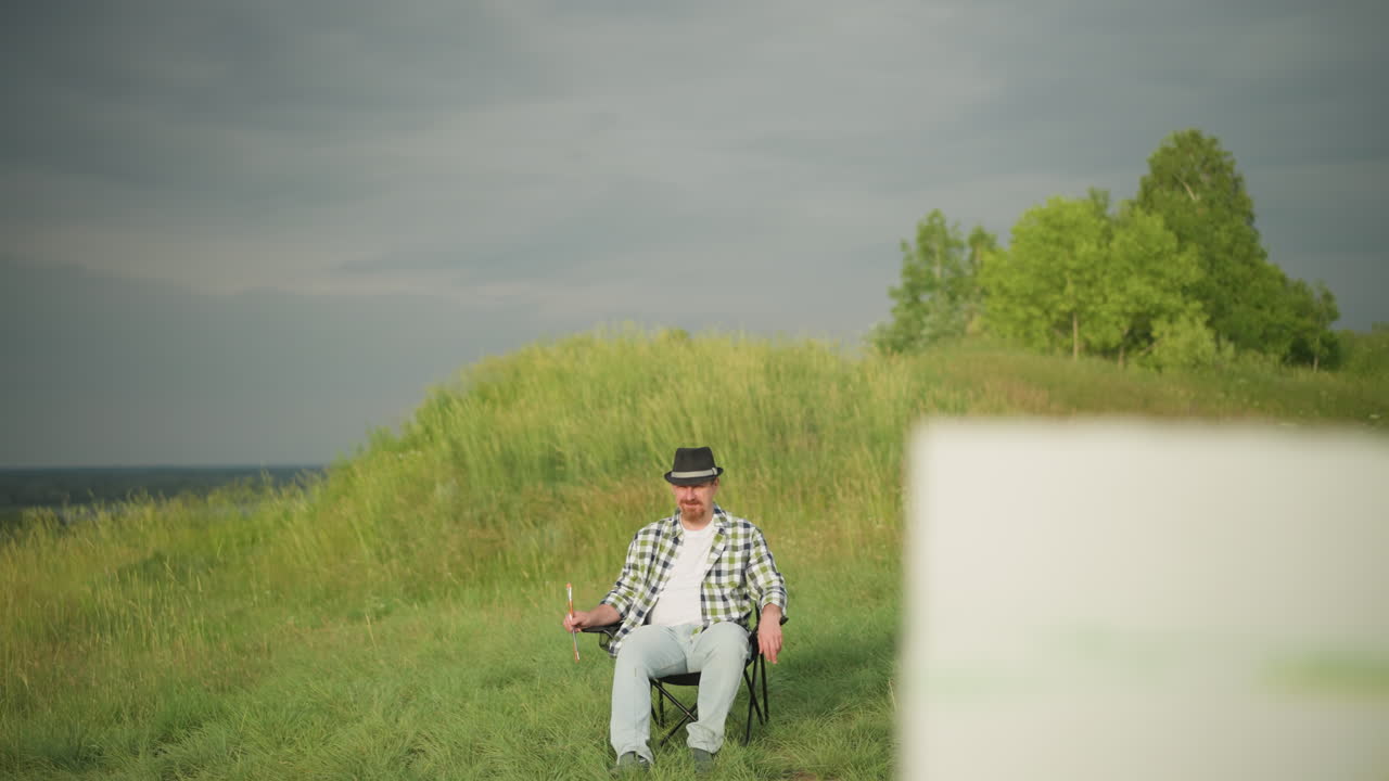 검은 모자, 체크 셔츠, 청바지를 입은 남자가 빛이 내리는 잔디에 있는 의자에 편안하게 앉아서 페인트 브러쉬를 들고 있다.