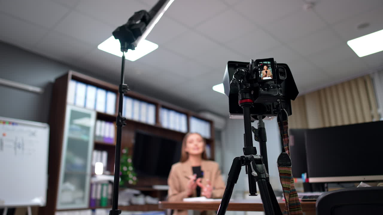 Camera on tripod and day-light lamp set in the office. Equipment records a female blogger talking at desk. Low angle view. Blurred backdrop.