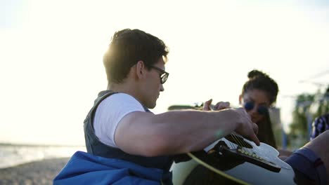 Young-man-playing-guitar-among-group-of-friends-sitting-on-easychairs-on-the-beach-and-singing-on-a-summer-evening-during-a-sunset.-Slowmotion-shot