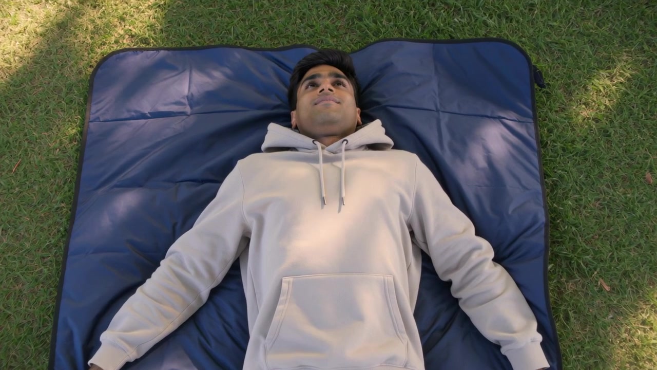 Pria Berhoodie Mengangkat Kepala ke Arah Matahari, Sedikit Berbalik di Atas Matras Biru di Rumput, dengan Bayangan Pohon