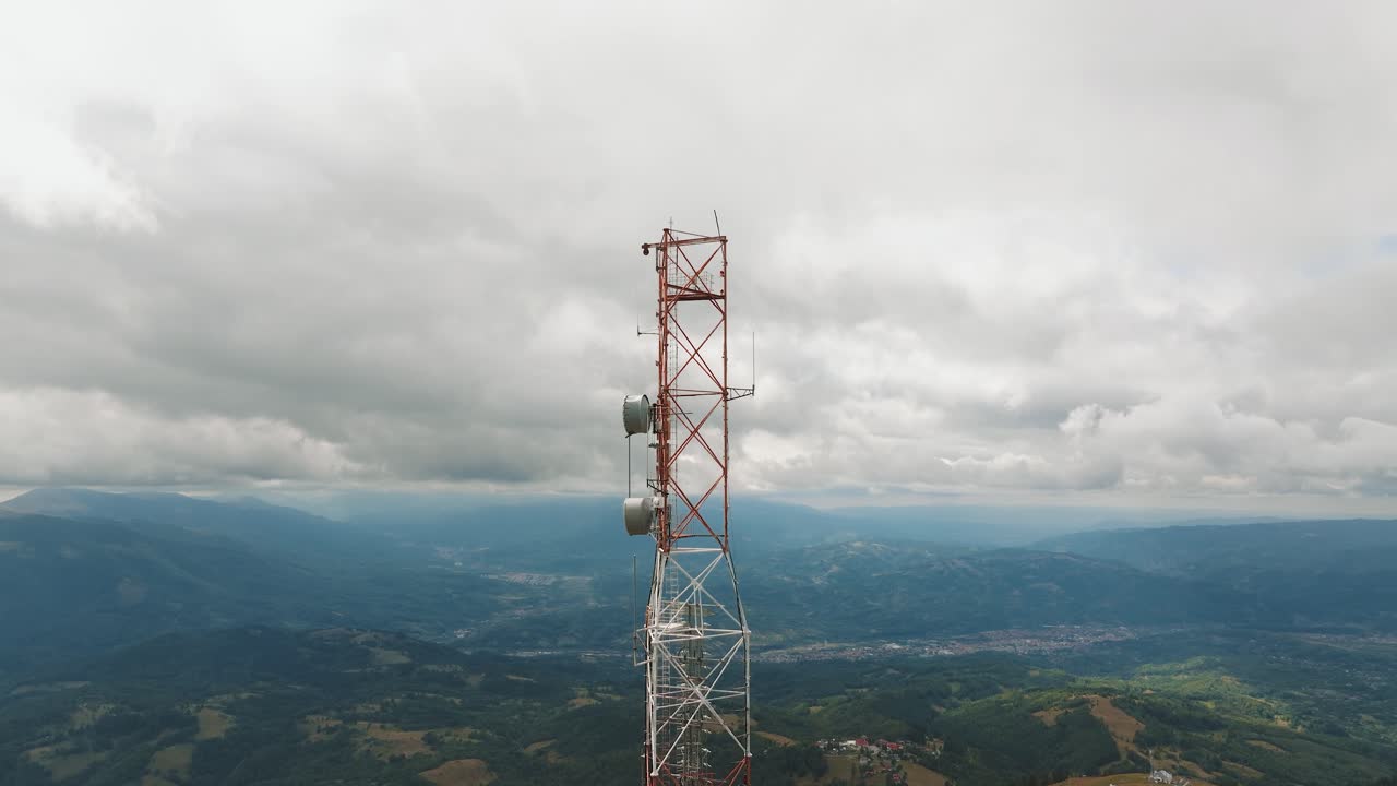 High altitude telecommunication tower holding multiple antennas, over a mountain landscape with distant small villages, aerial view