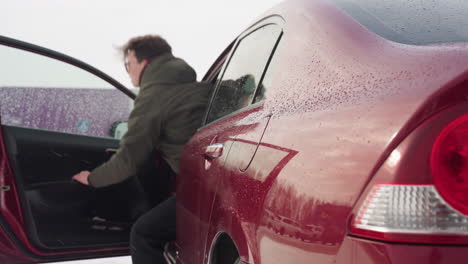 rear side view of red car with water droplets as businessman in winter jacket exits vehicle and shuts door, surrounding buildings and parked cars reflected clearly on glossy exterior in snowy setting