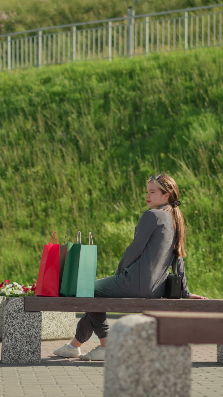 한 여인이 벤치에 다가와, 그 위에 다채로운 쇼핑 가방을 놓고, 앉아서, 왼쪽 다리를 앞으로 움직이면서 왼쪽 손을 편안하게 하고, 뒷면에 초록색 언덕이 있는 먼 곳을 쳐다본다.