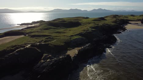 Aerial-view-Ynys-Llanddwyn-Welsh-island-with-shimmering-ocean-and-misty-Snowdonia-mountain-range-across-the-sunrise-skyline,-slow-reverse-shot