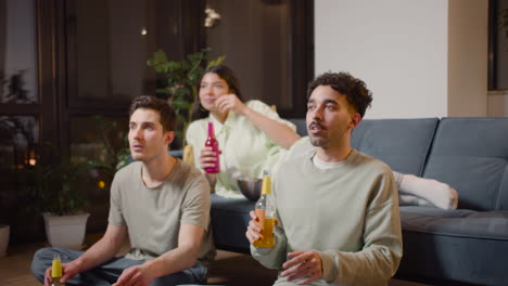 three friends watching comic movie on television sitting on couch and floor and drinking soda