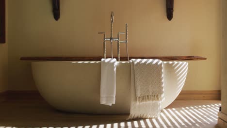 Oval-white-bathtub-in-minimal-modern-bathroom,-with-lines-of-sunlight-through-venetian-blind