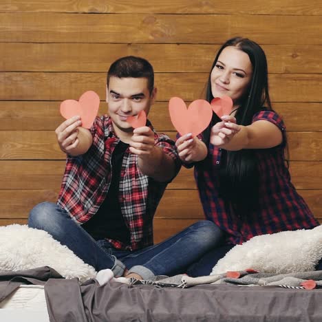 beautiful couple are sitting on the bed and showing red hearts by handmade in their hands on valentine's day