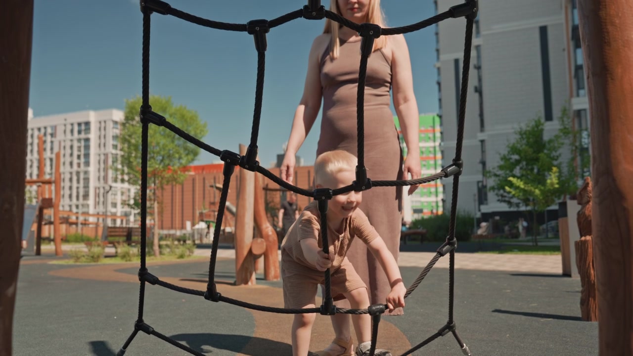 Young boy in brown outfit carefully navigates through black rope netting at outdoor playground with his mother standing nearby to assist. scene takes place on bright sunny day under clear