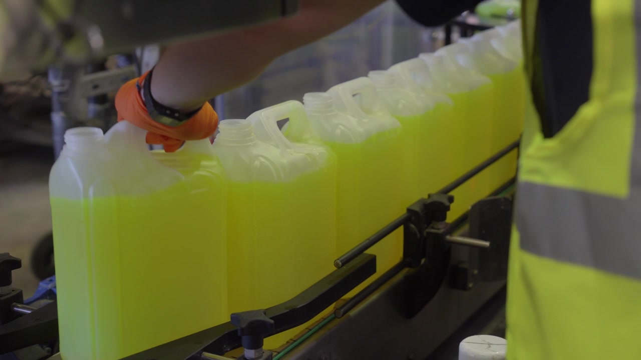 Factory worker in a safety vest inspecting neon yellow liquid canisters on a conveyor belt. Manual quality control in a chemical bottling plant.