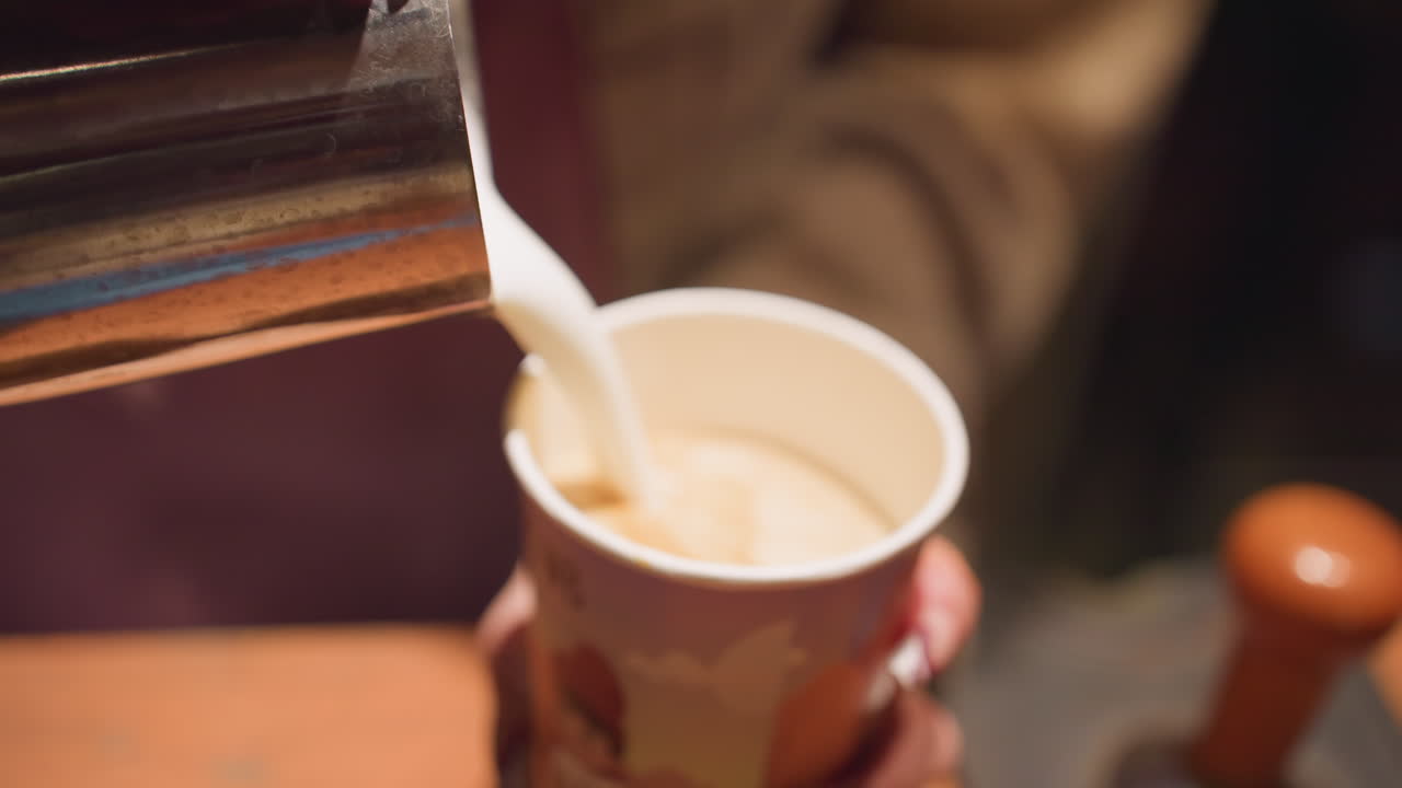 Close up of individual carefully pouring steamed milk from stainless jug into colorful coffee paper cup held by hand, preparing hot drink in cozy cafe setting with warm lighting