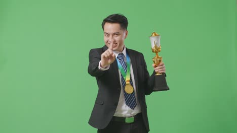 hombre de negocios asiático en traje y corbata con una medalla de oro y trofeo desaprobando sin señal del dedo índice y sonriendo a la cámara en el fondo de la pantalla verde en el estudio