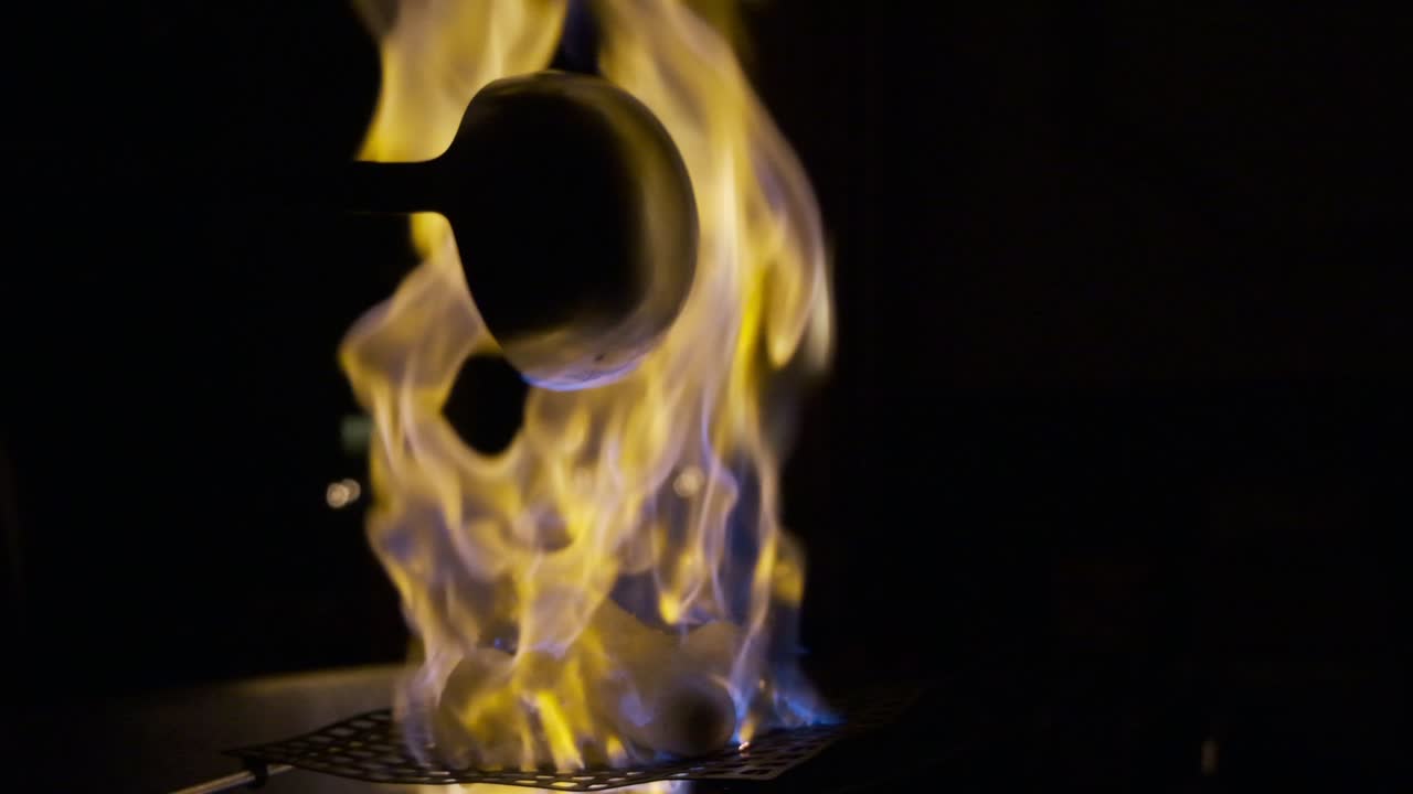 120fps super slow motion. Dark, illuminated by fire, german burnt punch prepared with burning rum poured from ladle over sugar into a big metal pot creating flames. Camera is close to the fire