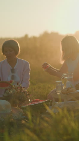 dos hermanas disfrutando de un picnic de verano con sandía fresca mientras una recoge semillas y la otra toma un bocado, a su lado, una joven está inclinada, saboreando su rebanada