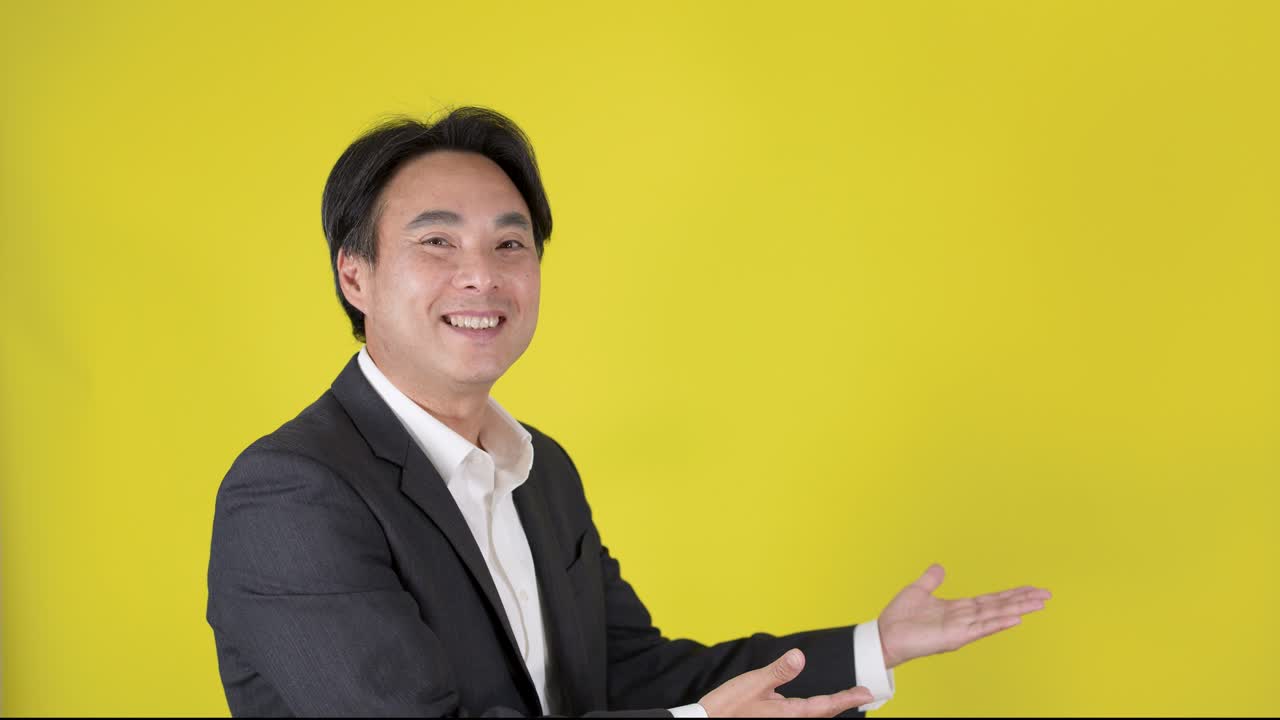 Smiling businessman in suit points and gestures confidently, bright studio lighting, static camera, yellow backdrop