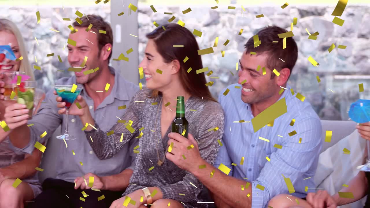 Sei adulti seduti su un divano con coriandoli dorati che cadono mentre il gruppo alza i bicchieri brindando per celebrare un evento.