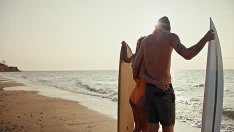 Rear-view-of-a-happy-blond-guy-in-a-hat-with-a-bare-torso-and-in-dark-green-shorts-stands-with-his-blonde-girlfriend-in-an-orange-swimsuit,-they-hold-surfboards-standing-on-a-sandy-beach-and-look-into-the-sea-at-Sunrise-in-the-morning