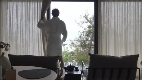 Male-Guest-In-White-Bathrobe-Opens-Curtains-In-Hotel-Room,-Exposing-Scenic-Nature-Outside-Glass-Door