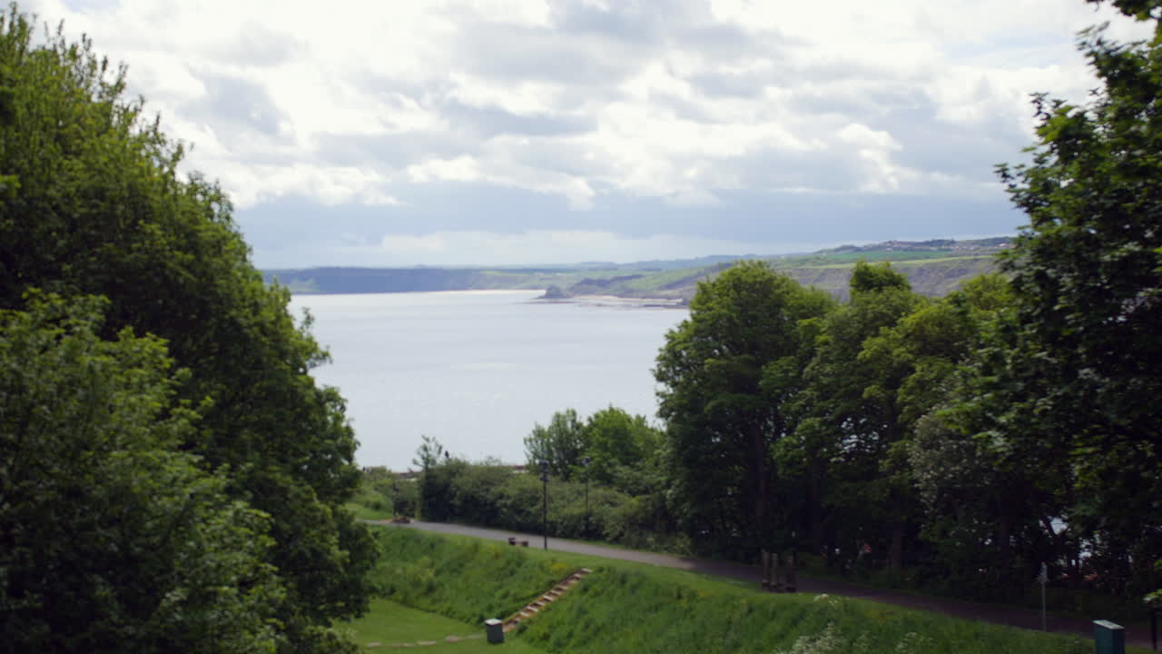 A coastal landscape view from Scarborough, North Yorkshire, England shows the sea beyond green trees, with cliffs and distant shoreline visible across the bay
