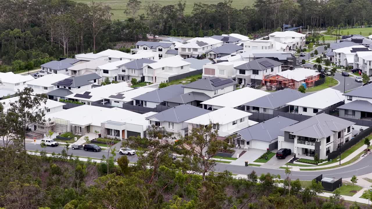 Smooth aerial camera movement reveals contemporary suburban housing, lush greenery, and mountain backdrop in daylight