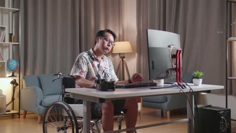 hombre asiático cansado en silla de ruedas sacudiendo la cabeza y teniendo dolor de cabeza mientras edita el video por un escritorio al lado de la cámara en casa