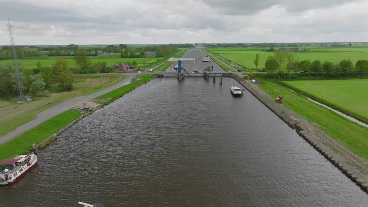 A loaded cargo barge approaches a bascule bridge on a Dutch canal; banks with parked boats and service track alongside