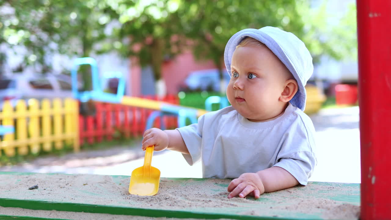अपने सिर पर पनामा टोपी पहने नीली आँखों वाला एक छोटा लड़का रेत में खेल रहा है। बच्चे धूप वाले दिन सड़क पर रेत से खेलते हैं।