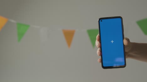 teléfono móvil de pantalla azul contra banderines en colores de la bandera nacional irlandesa colgado en el estudio para celebrar el día 2 de san patricio