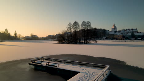 Aerial-flyover-jetty-and-frozen-lake-covered-with-white-snow-during-sunset---Beautiful-old-historic-city-with-church-in-background---Poland,Europe